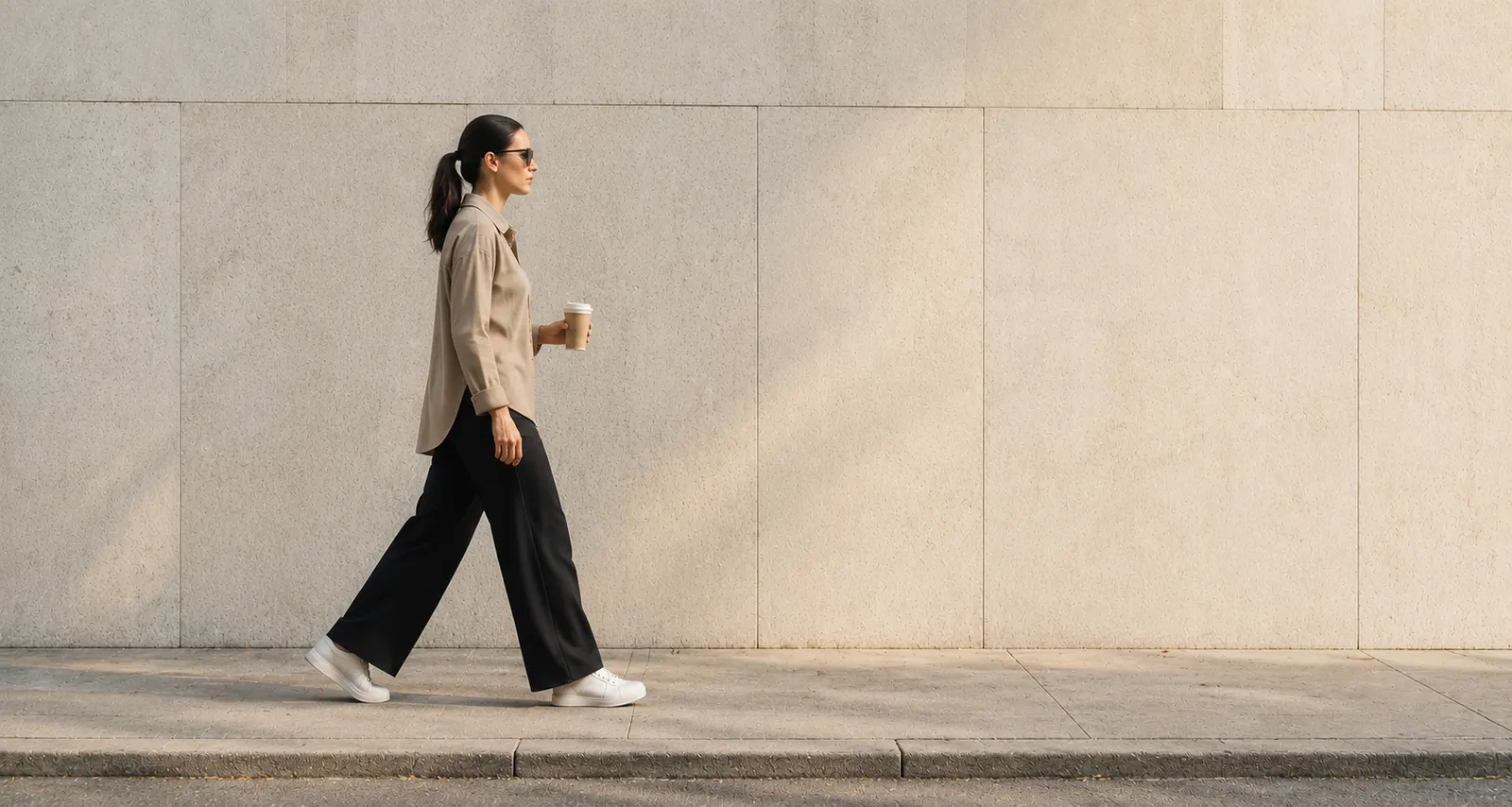 Business owner walking past a limestone wall with a coffee cup, moving her luxury trade business forward with PAIWE's liquidity network.